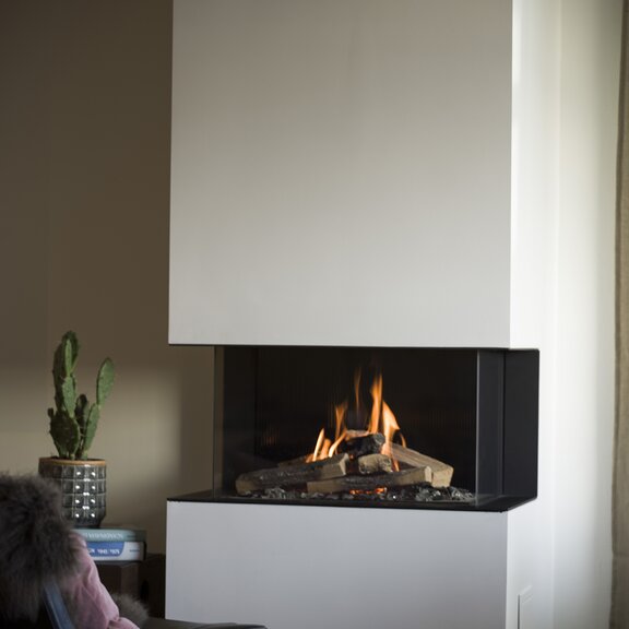 Cheminée à gaz VISIO 90 3S en tant que cheminée tri-face avec une flamme de gaz calme dans un habillage blanc à côté d'une plante et d'un fauteuil de lecture