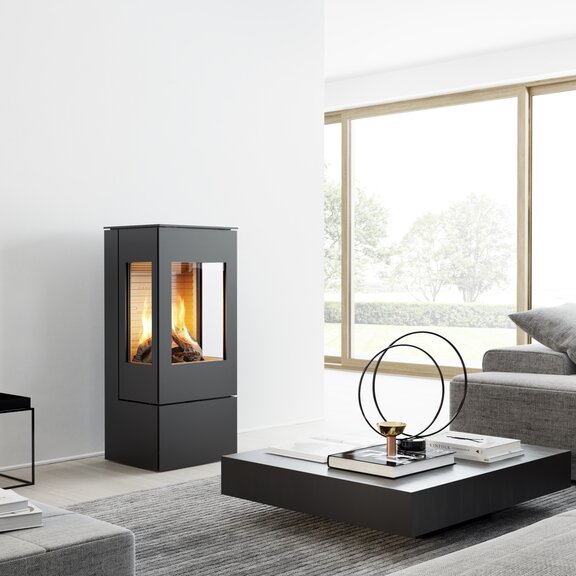 Poêle à gaz NEXO 100 en noir avec porte en acier et deux vitres latérales dans un salon moderne et lumineux